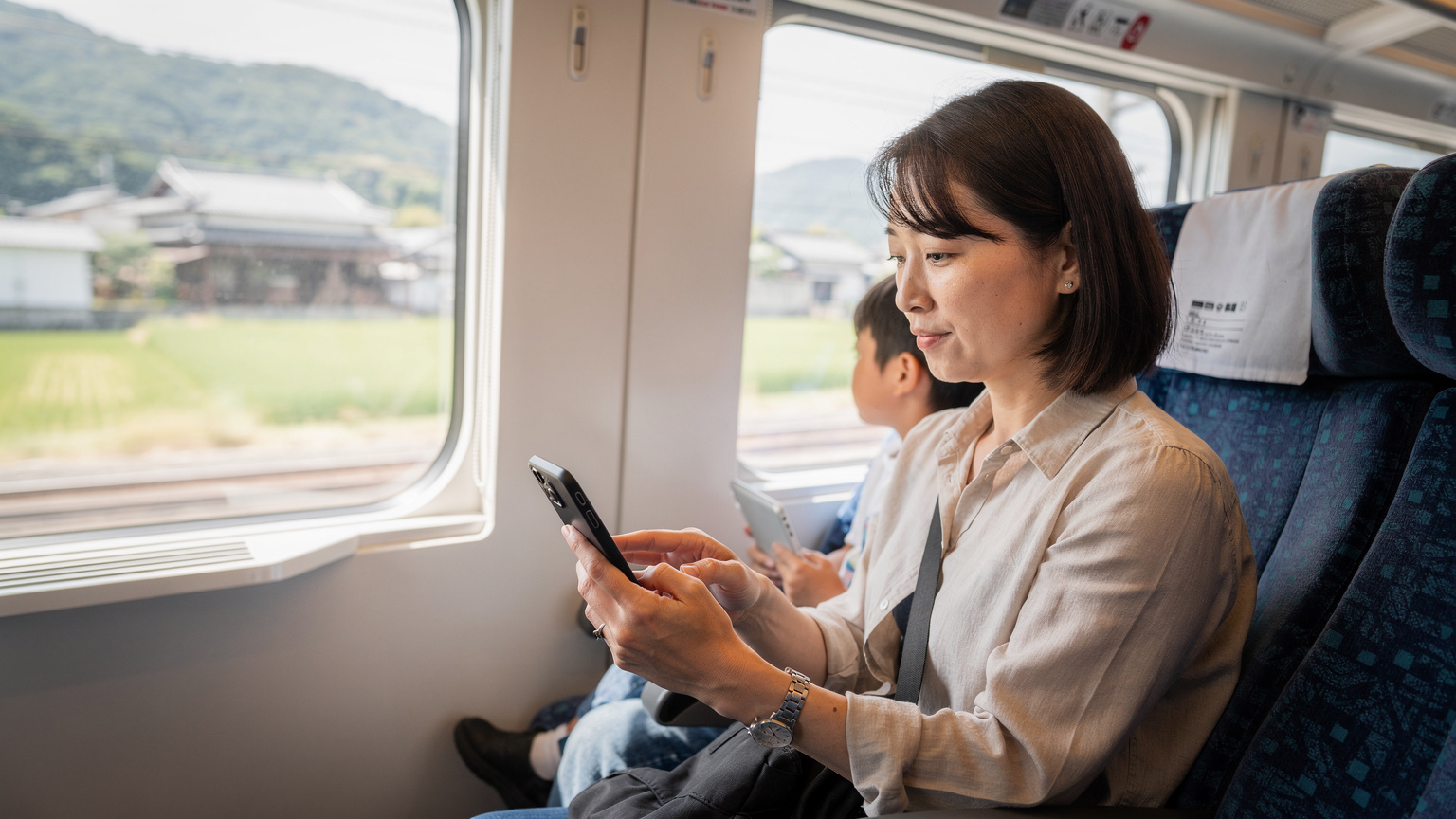 Eine Mutter checkt auf ihrem Smartphone die eSIM-Einstellungen, während ihr Kind im Hintergrund in einem japanischen Zug aus dem Fenster blickt und ein Tablet hält.