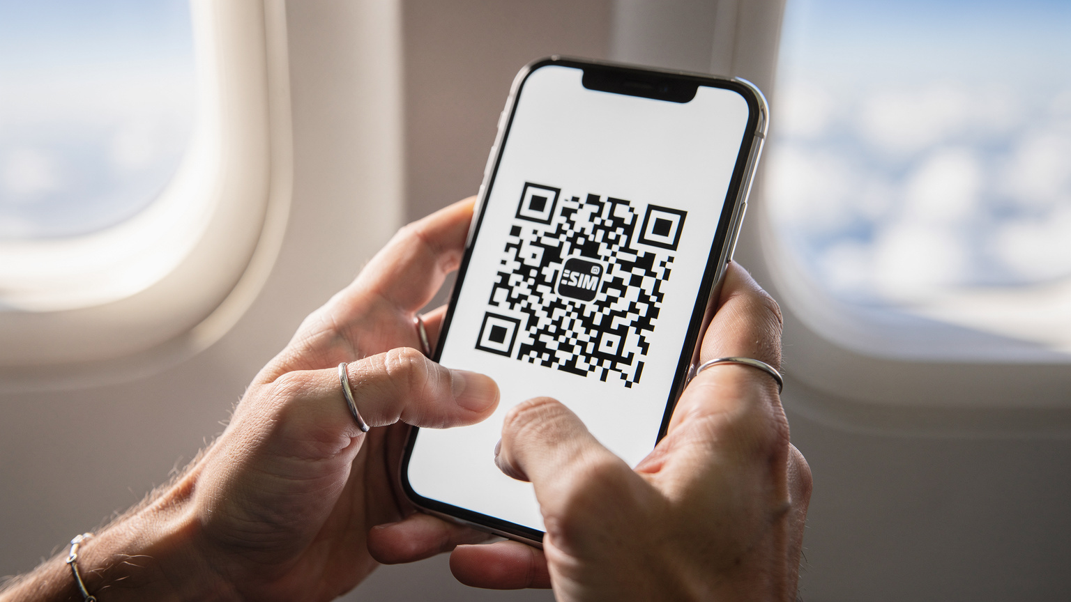 A close-up of a person's hands holding a smartphone, with the screen displaying an eSIM activation QR code, against a blurred backdrop of an airplane window at cruising altitude.